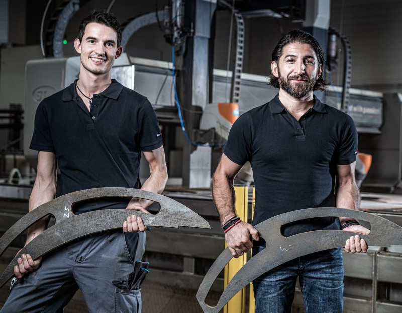 Zwei Eisen Neumüller Mitarbeiter halten von Wasserstrahlschneidanlagen ausgeschnittene Stahlteile 